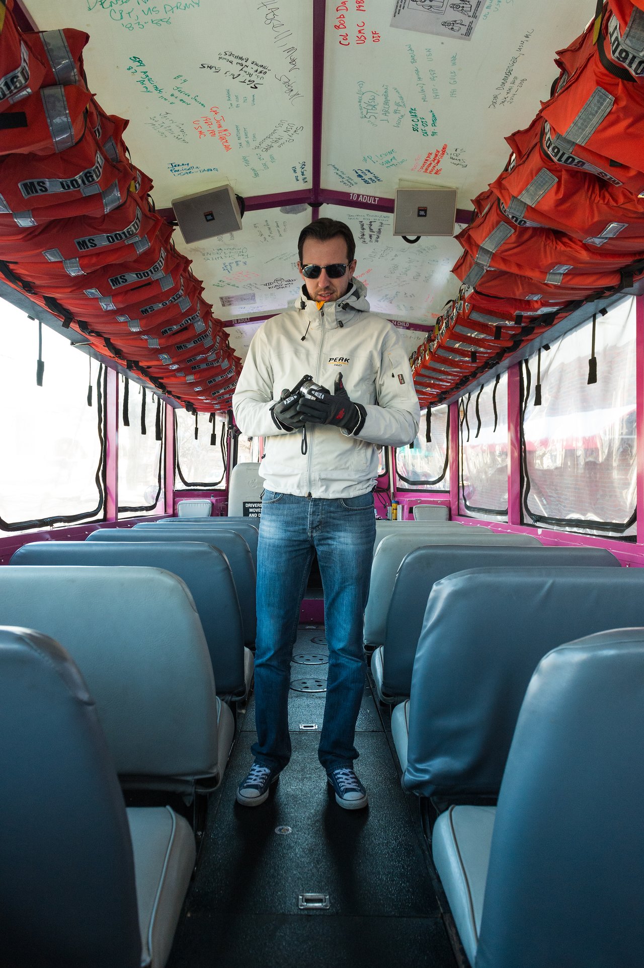 A man wearing sunglasses and gloves stands inside a Boston Duck Tour vehicle, looking down at his hands.