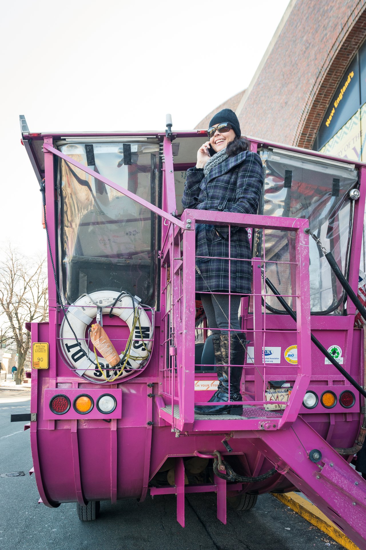 A person stands on the back of a pink Boston Duck Tour vehicle, talking on the phone and smiling.