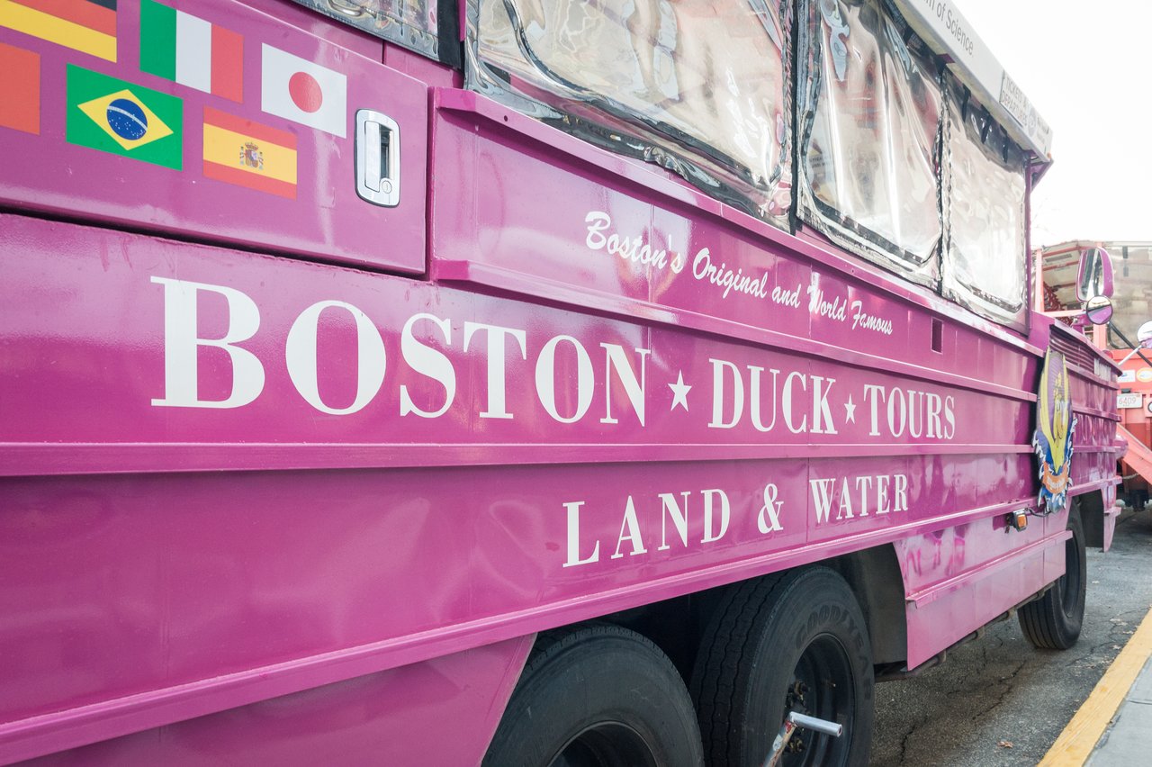 A pink Boston Duck Tours vehicle parked on the street, featuring large text and international flags on its side.