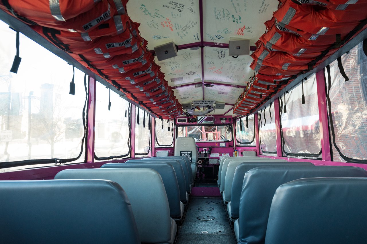 The interior of a Boston Duck Tour vehicle with rows of blue seats, life jackets overhead, and a clear plastic enclosure.
