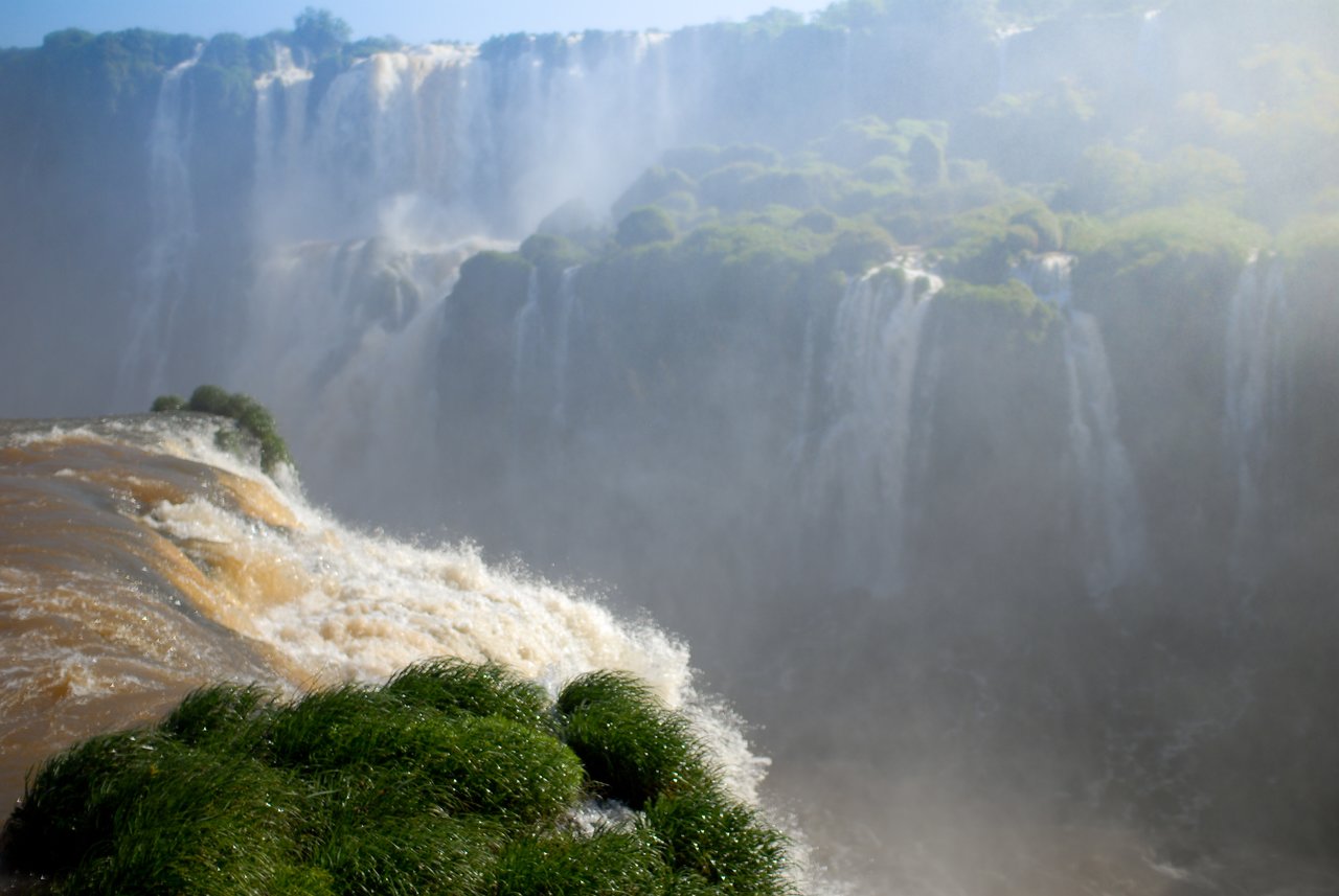 Water cascades over the edge of Iguazu Falls, with mist rising and green vegetation covering nearby rocks.