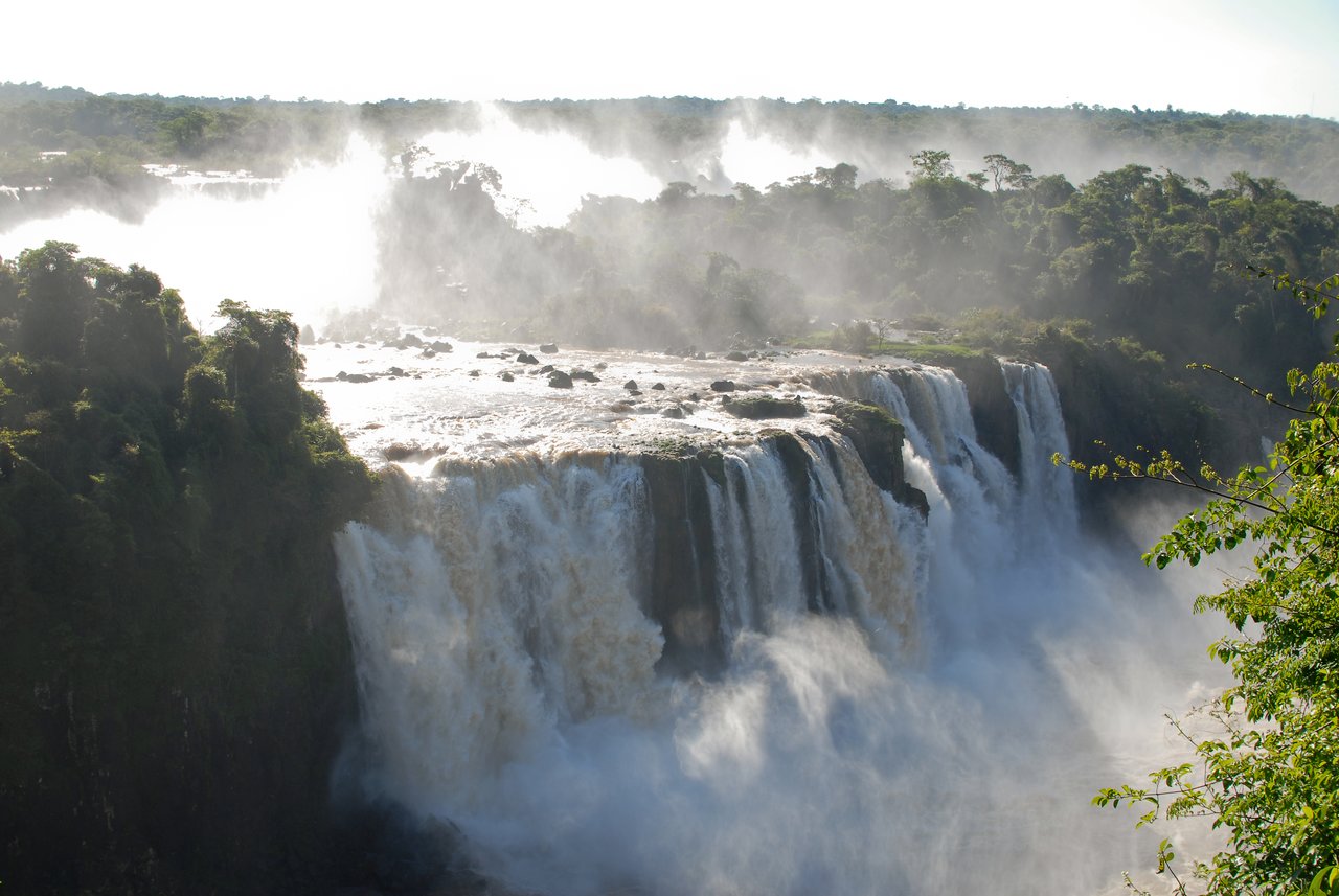 Large waterfalls cascade over a rocky edge, creating mist and turbulence, surrounded by green vegetation.