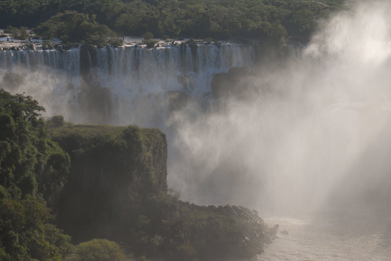 Water cascades down Iguazu Falls, creating mist that rises above the river, surrounded by green cliffs and trees.