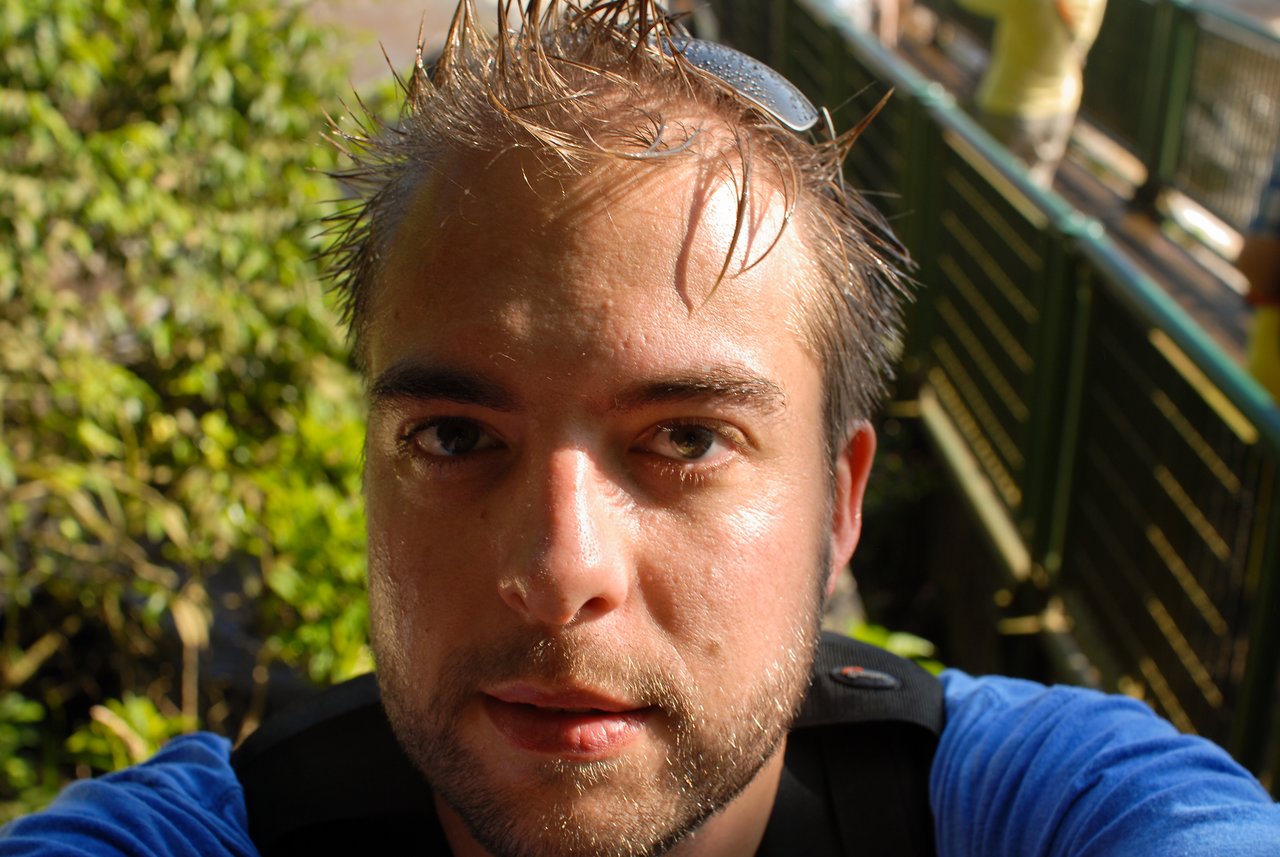 A person with wet hair and sunglasses on their head takes a close-up selfie on a walkway near greenery.