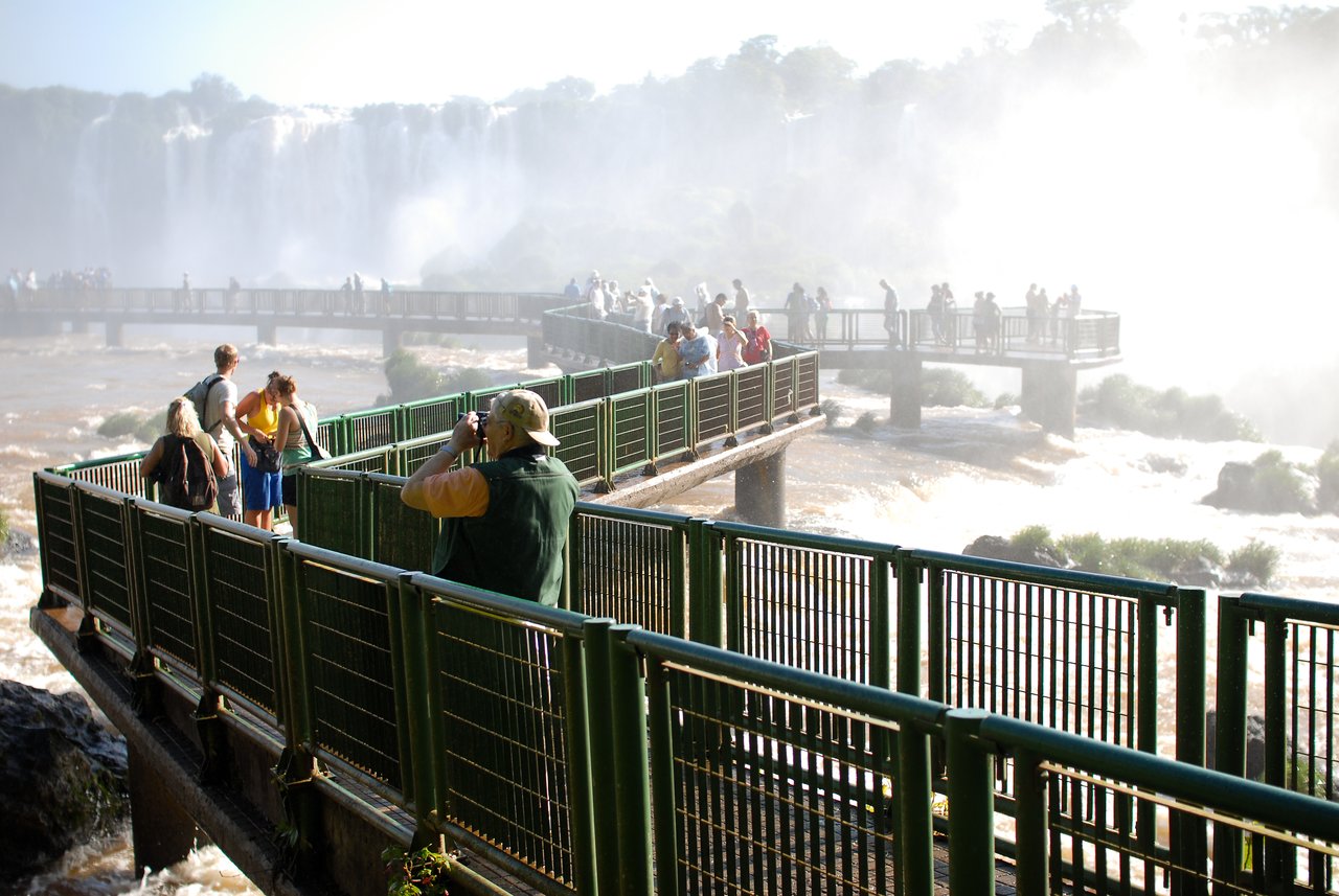 Tourists walk on a metal walkway near Iguazu Falls, taking photos and observing the rushing water and mist.