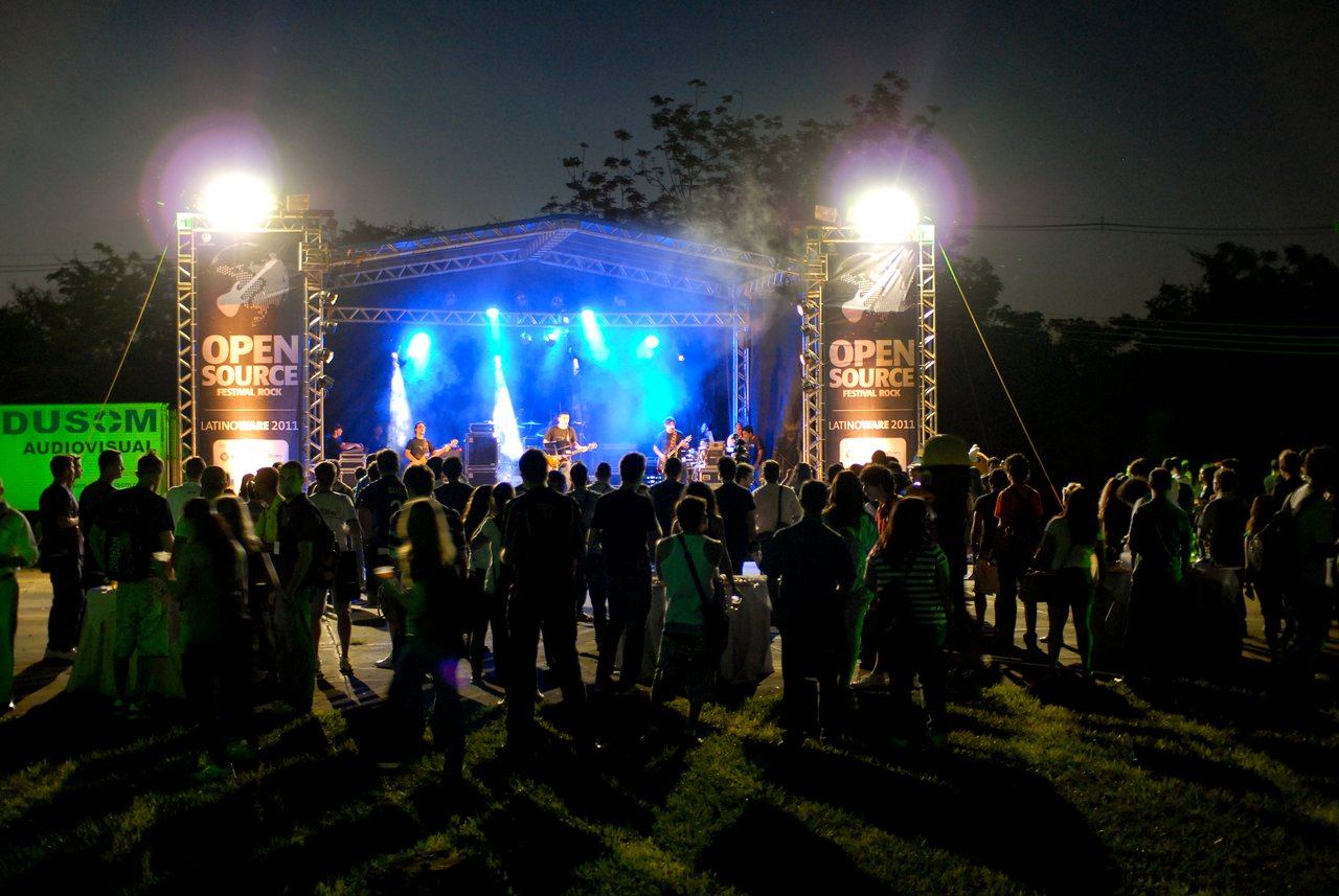 A live rock band performs on an outdoor stage at night, with a crowd gathered to watch.