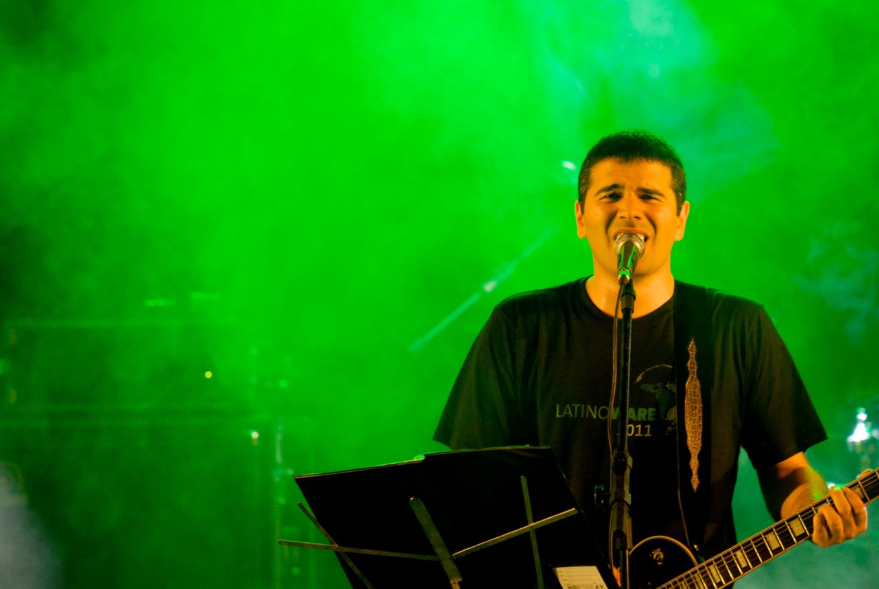 A musician sings into a microphone while playing an electric guitar on stage, with a music stand in front.