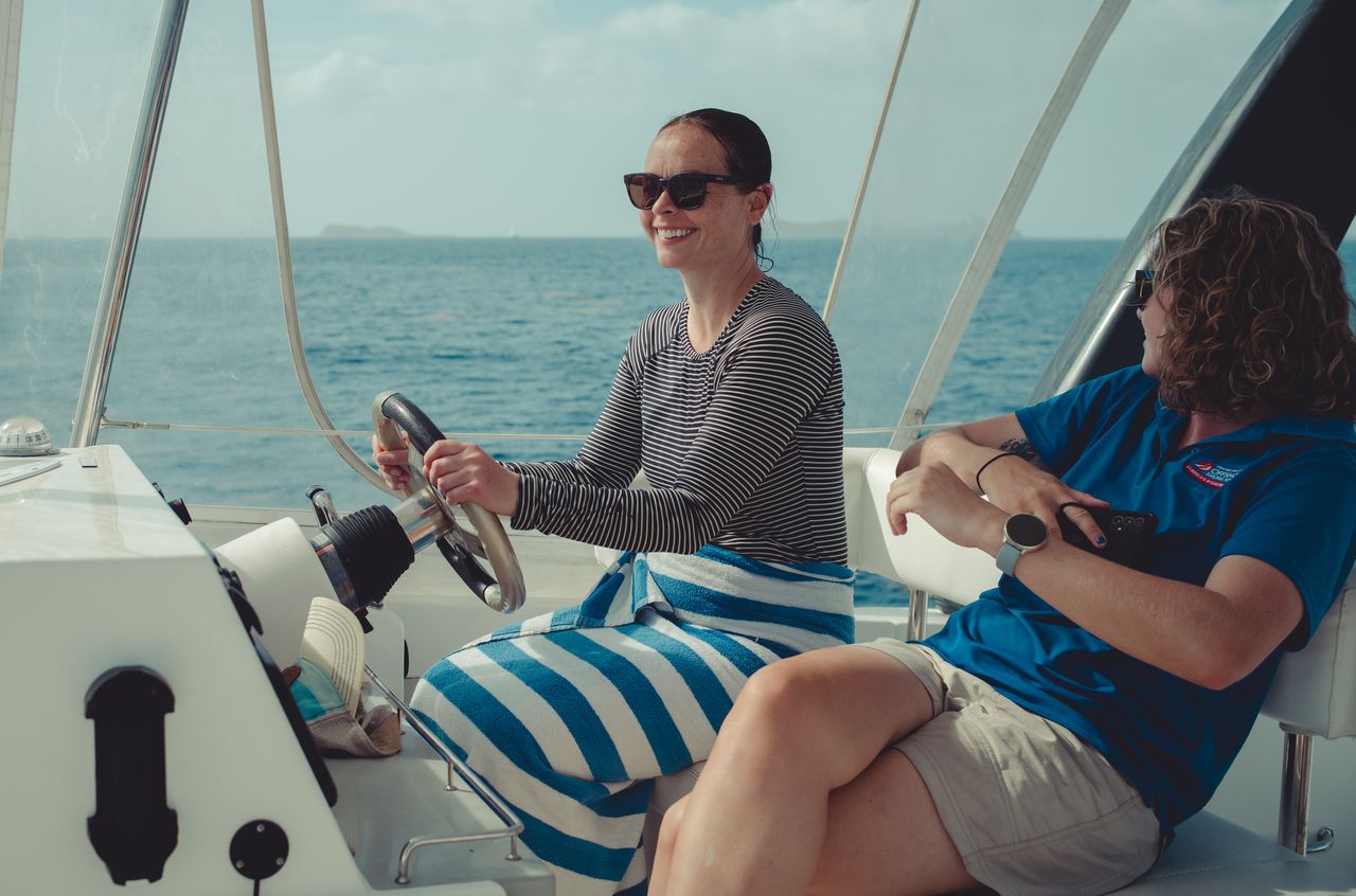 A woman smiles while steering a boat, with a passenger sitting beside her.