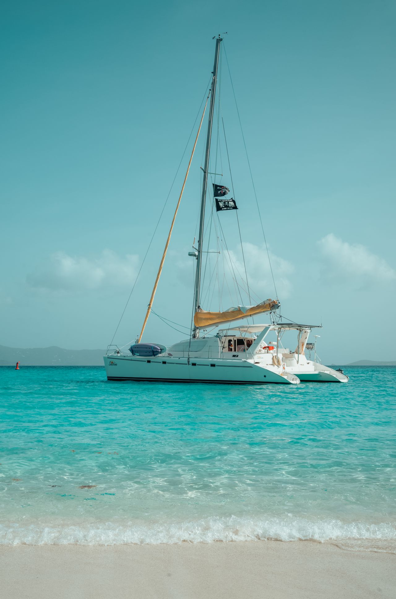 A white catamaran with a tall mast floats on clear turquoise water near a sandy beach.