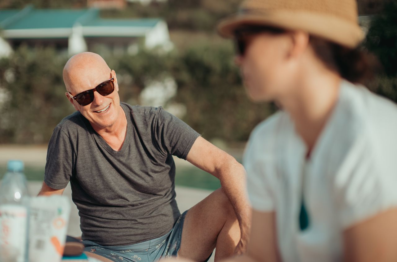 A man in sunglasses and a gray shirt smiles while chatting with a woman wearing a hat.