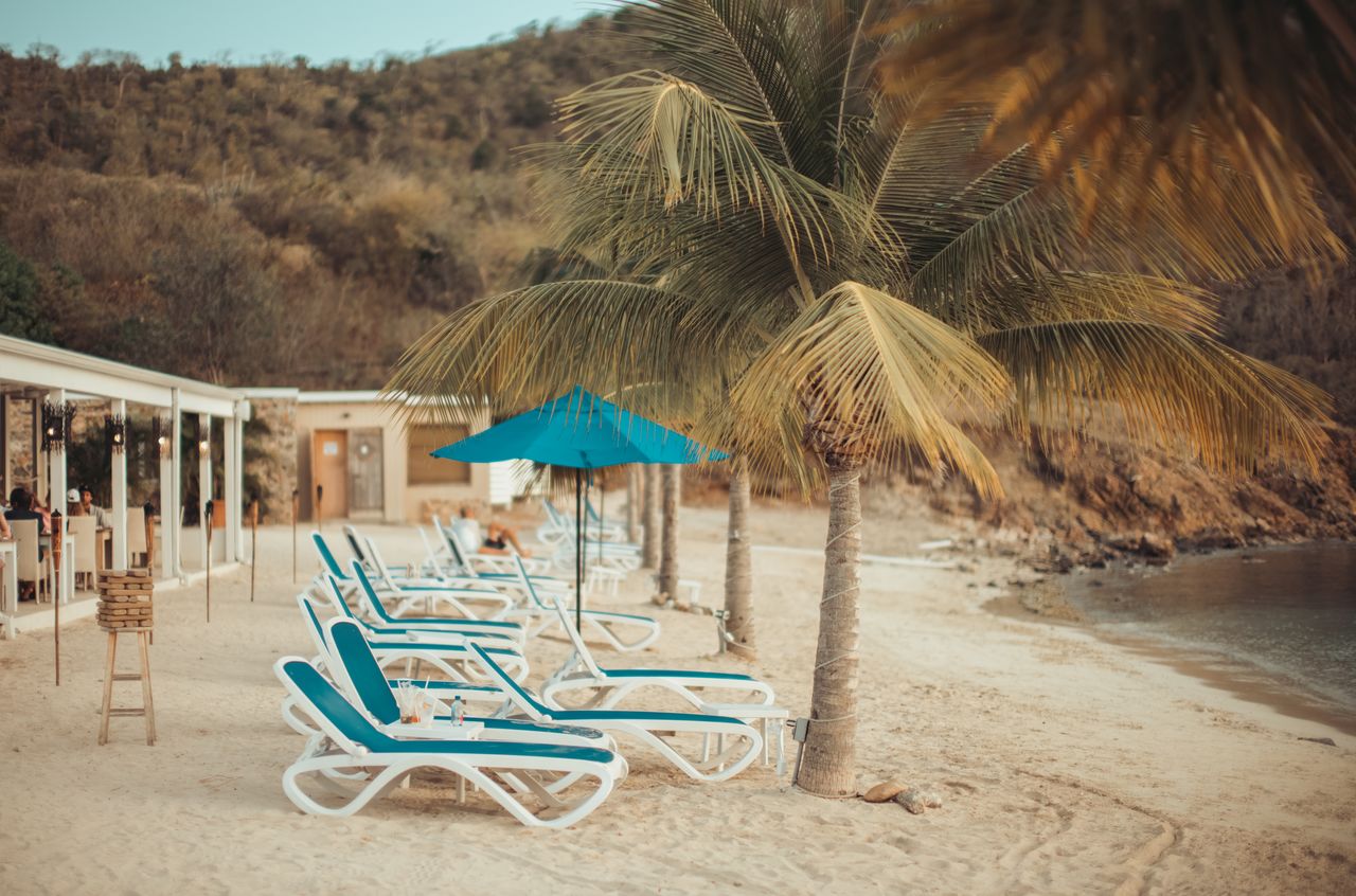 Lounge chairs with blue cushions and umbrellas on a sandy beach, with palm trees and a beachside restaurant nearby.