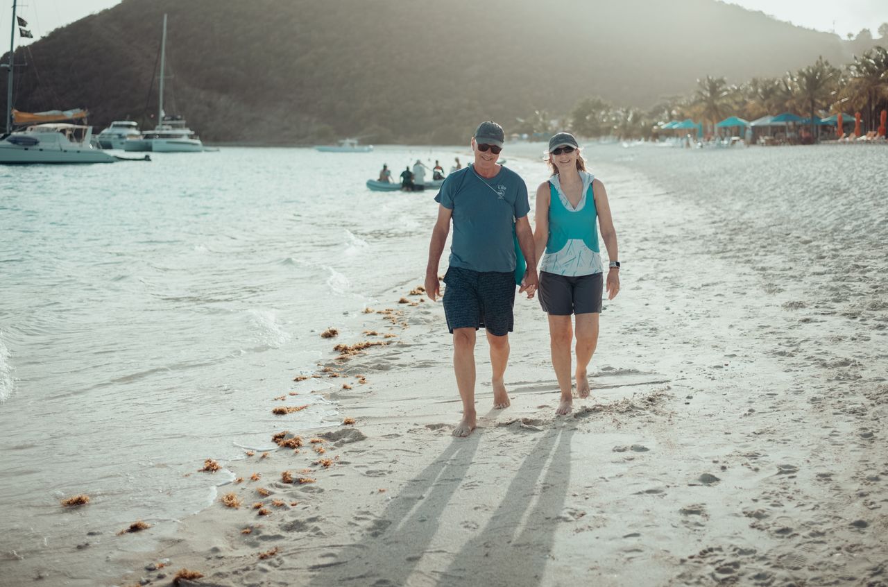 A couple walks barefoot on a sandy beach, holding hands, with boats and people in the water behind them.