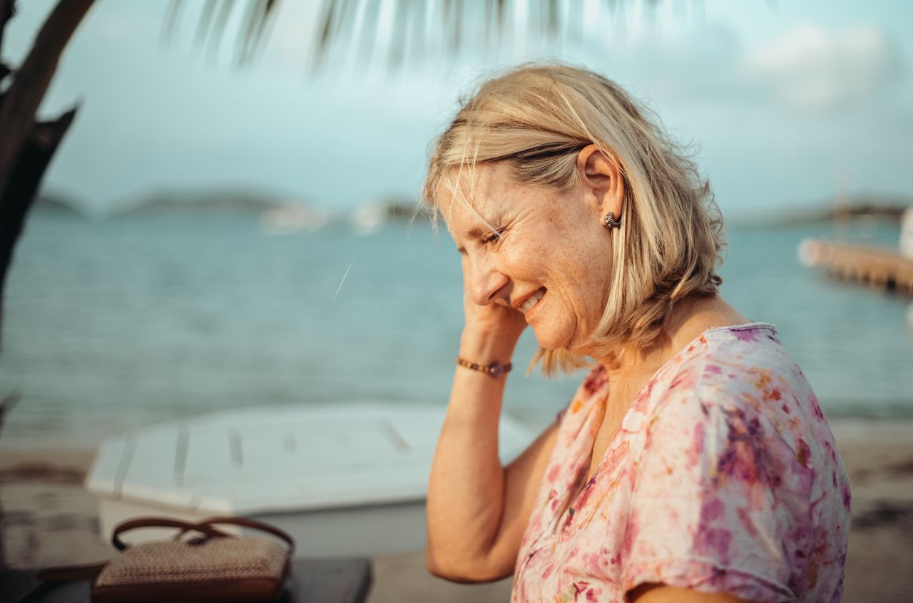 A woman with blonde hair smiles while touching her ear, standing near the water with boats in the background.