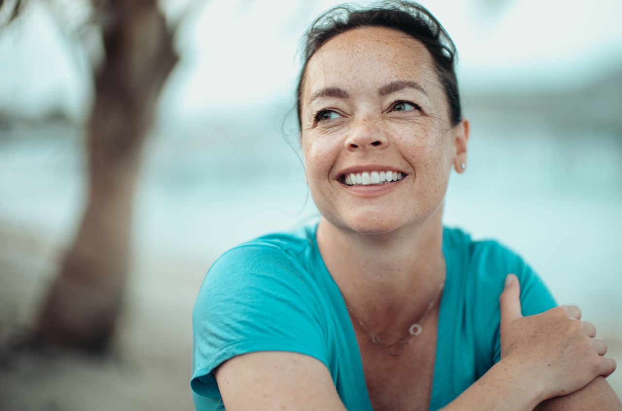 A woman in a turquoise shirt smiles while looking to the side, sitting outdoors with a blurred background.