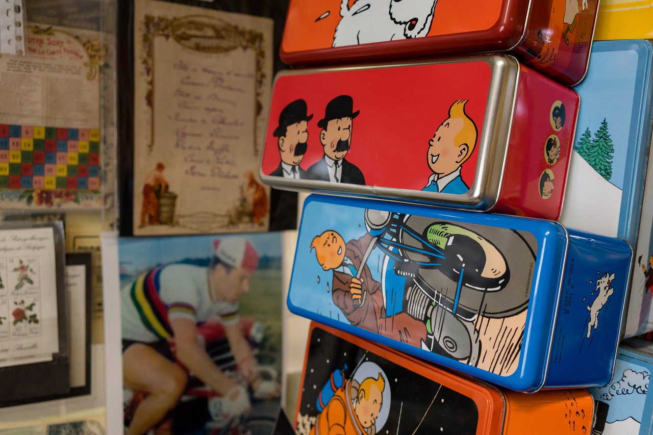 Stacked metal tins featuring Tintin comic illustrations, with vintage documents and a cyclist photo in the background.