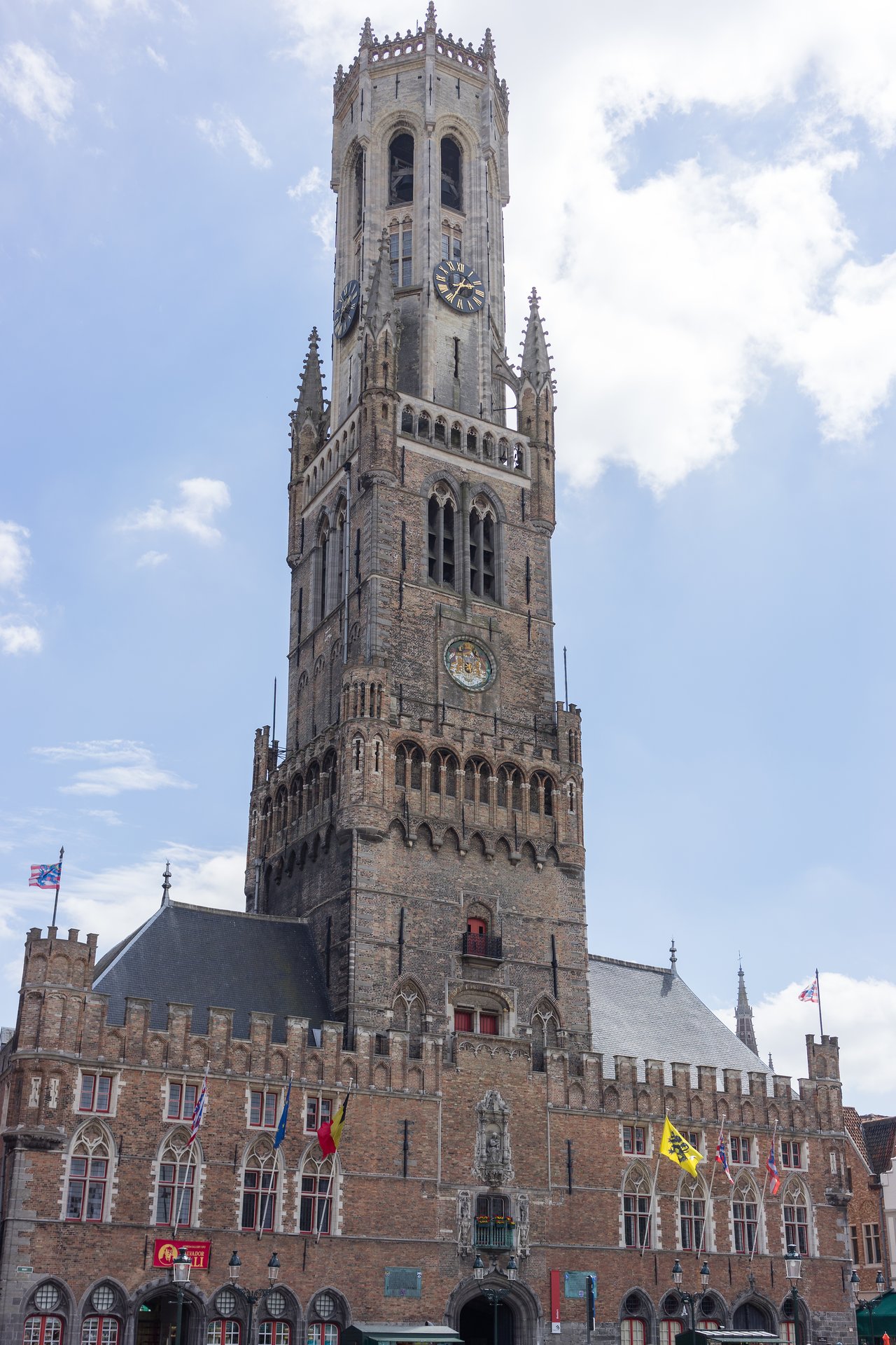 The Belfry of Bruges, a tall medieval bell tower with clocks, stands against a partly cloudy sky.