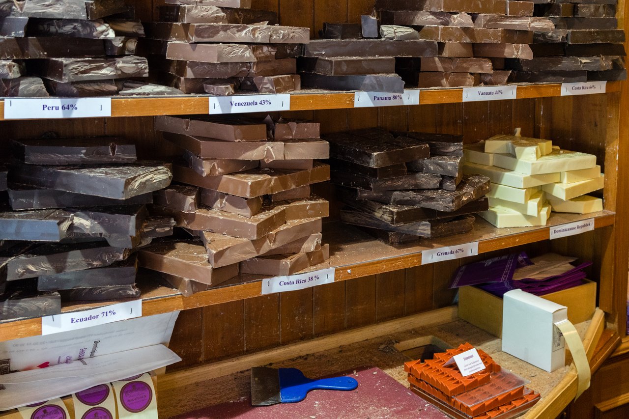 Stacks of dark, milk, and white chocolate bars labeled by cocoa origin on wooden shelves in a chocolate shop.