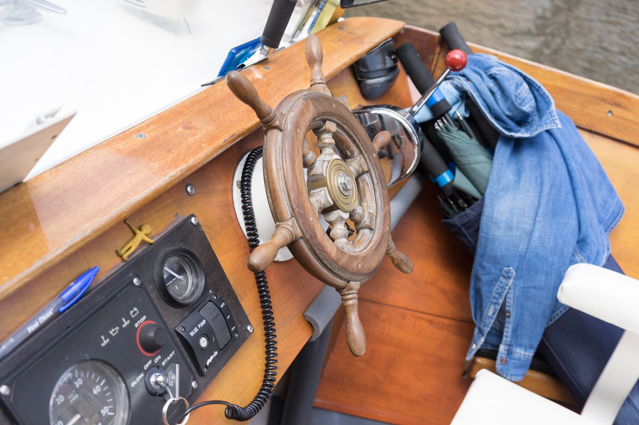 Wooden boat steering wheel and control panel with switches and gauges, alongside stored umbrellas and a denim jacket.