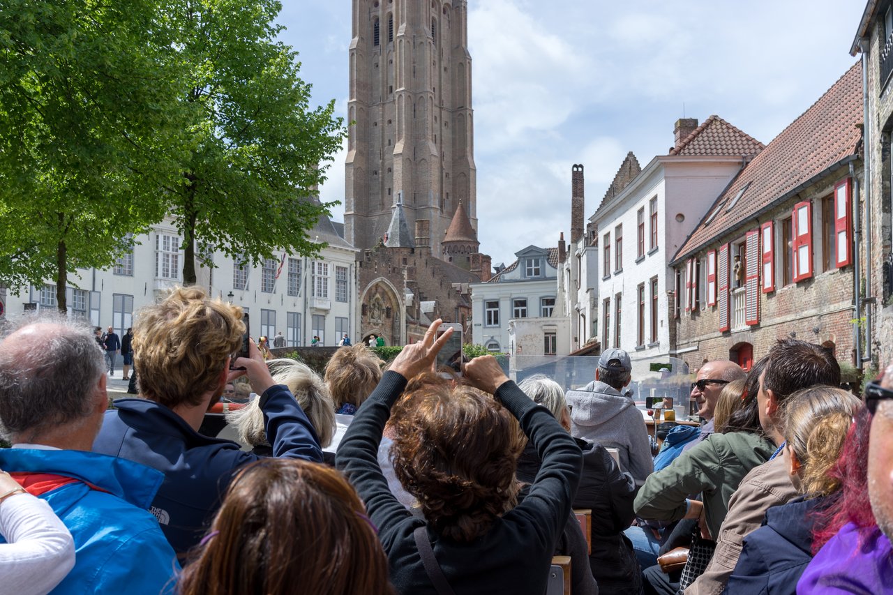 A group of tourists on a boat tour in Bruges, taking photos of a tall historic church.