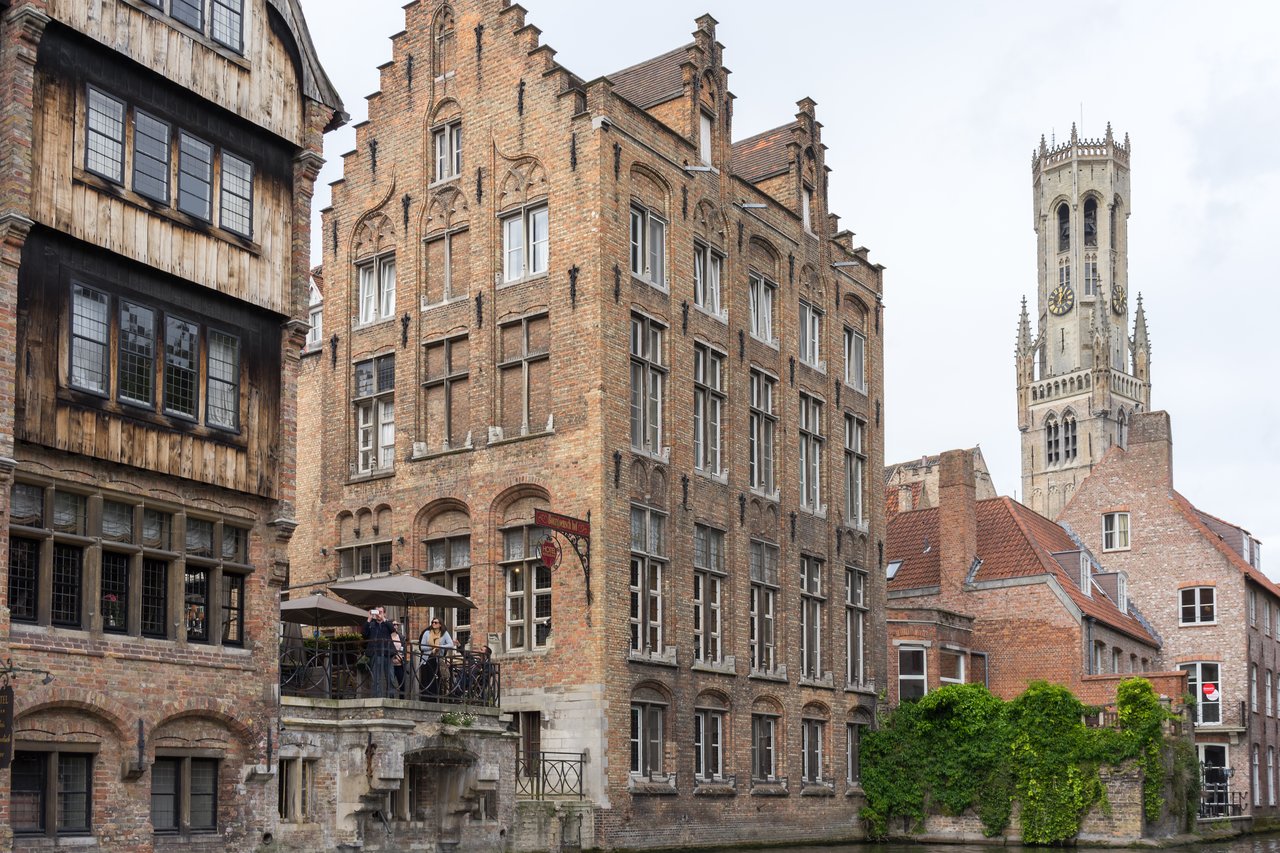 Historic brick buildings and a canal in Bruges, with people standing on a terrace.