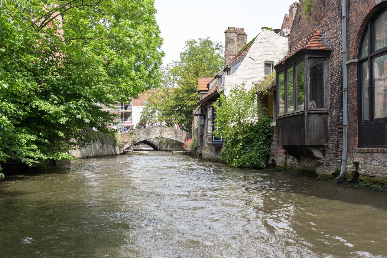 A canal view in Bruges with historic buildings, a stone bridge, and tourists standing on top.