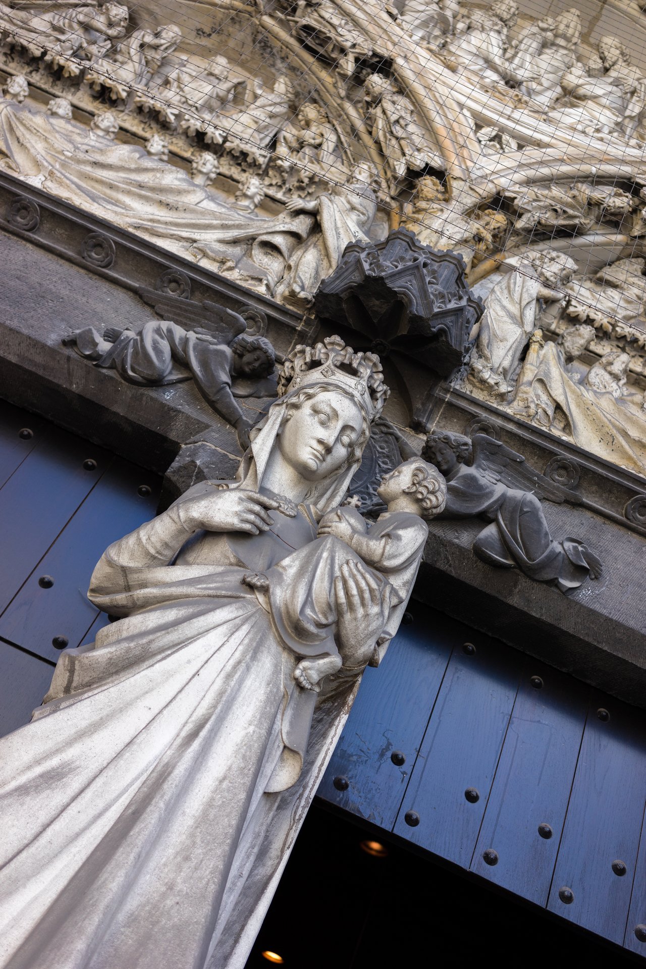 Stone statue of Mary holding baby Jesus, positioned above a doorway with detailed religious carvings in the background.