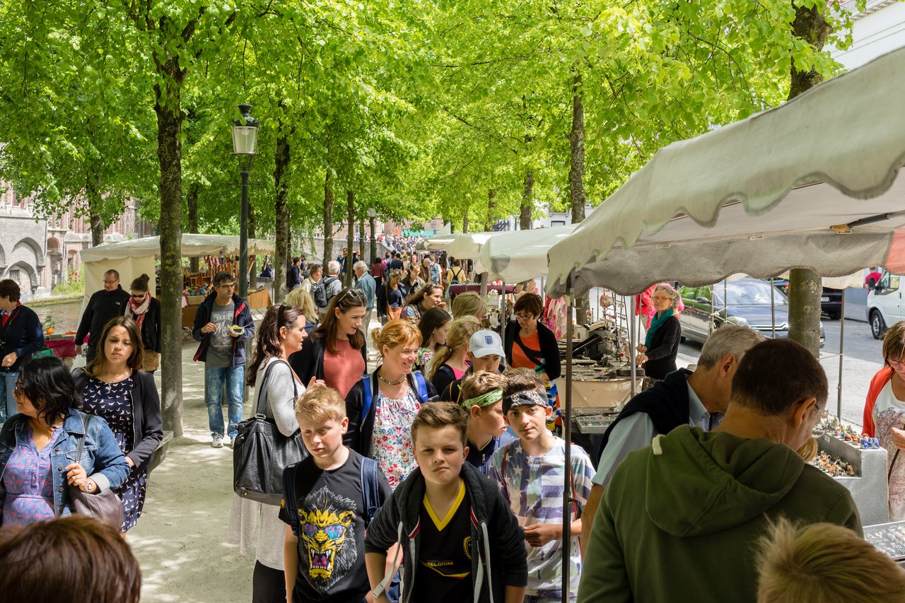 A busy outdoor market with people walking, browsing stalls, and shopping under white tents lined along a tree-covered path.