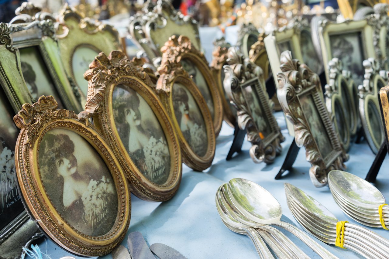 A market stall displaying ornate vintage photo frames with black-and-white portraits and stacks of silverware on a table.