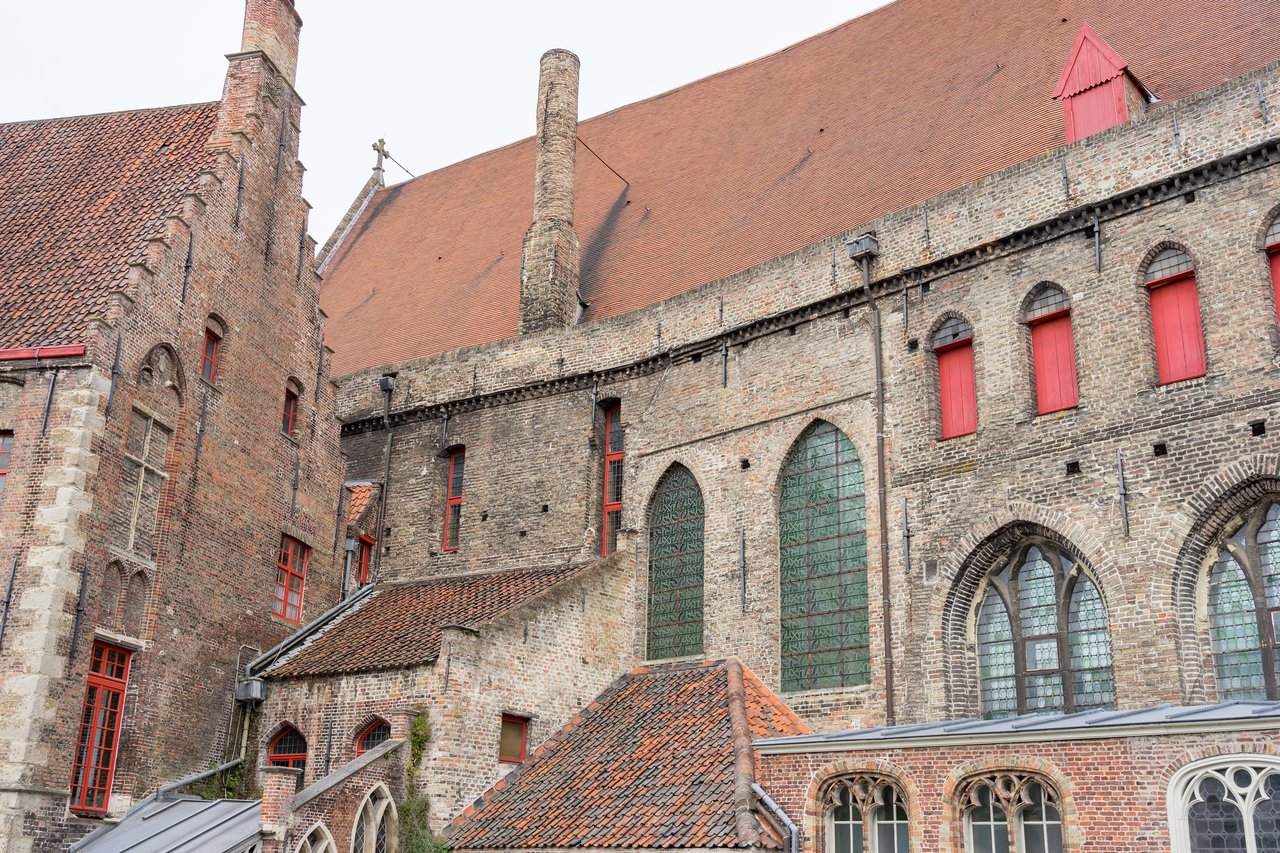 Old brick hospital building with arched windows, red shutters, and a steep roof in Bruges.