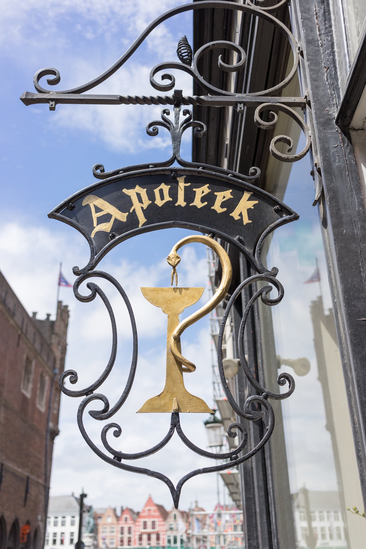 Ornate metal pharmacy sign with "Apotheek" in gold lettering and a golden bowl with a snake, hanging outside a building.