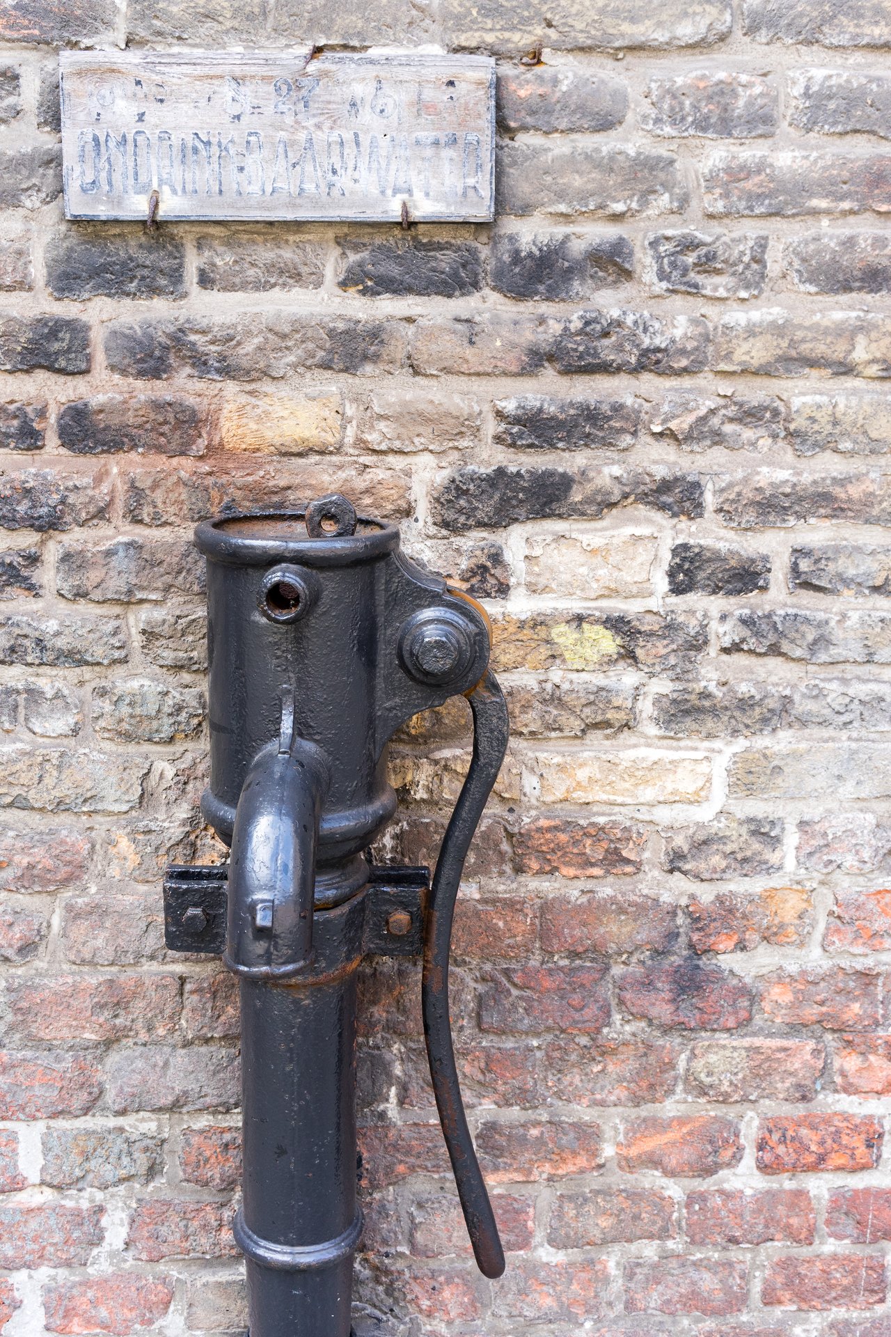 An old black metal water pump with a long handle, mounted on a brick wall with a worn sign above.