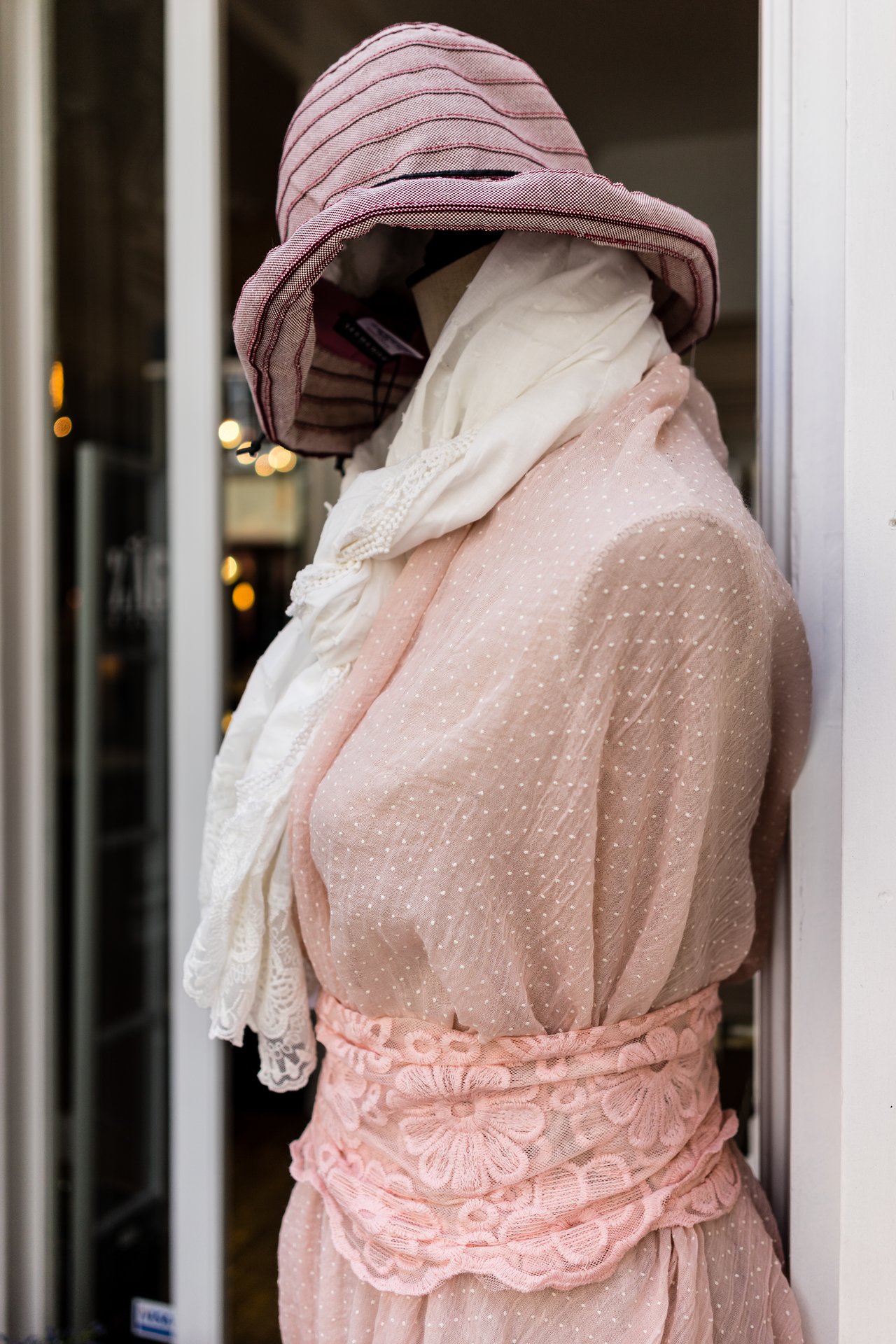 A mannequin dressed in a pink dress, white scarf, and hat is displayed outside a clothing store.