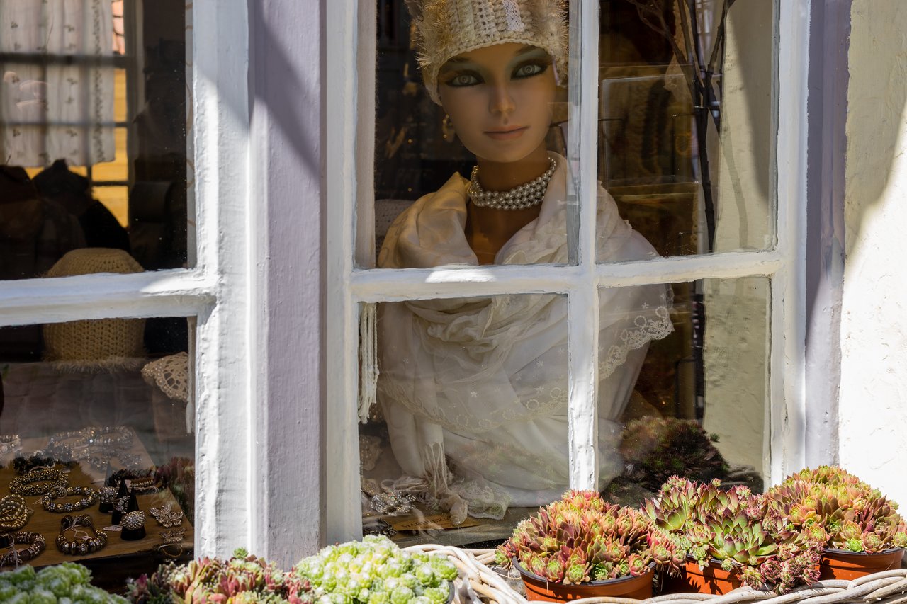 A mannequin dressed in white with a pearl necklace is displayed in a shop window, surrounded by jewelry and plants.