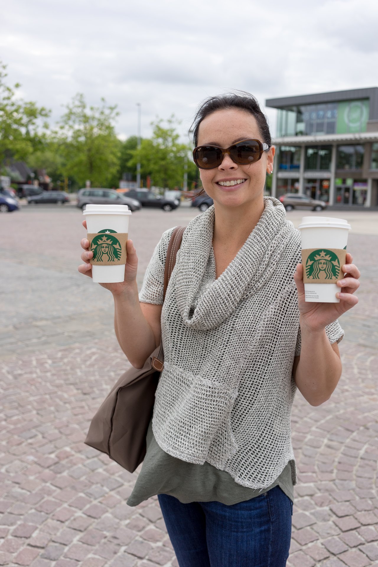 A smiling woman wearing sunglasses holds two Starbucks coffee cups while standing outdoors in a public square.