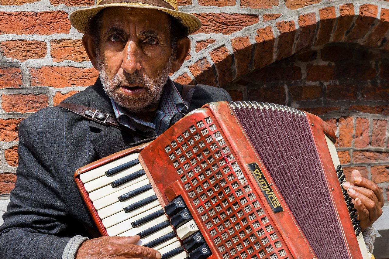 An elderly street artist wearing a hat plays an accordion against a brick wall.
