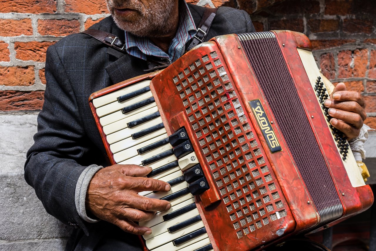 A street artist plays an accordion, pressing the keys and buttons while wearing a dark jacket against a brick wall.