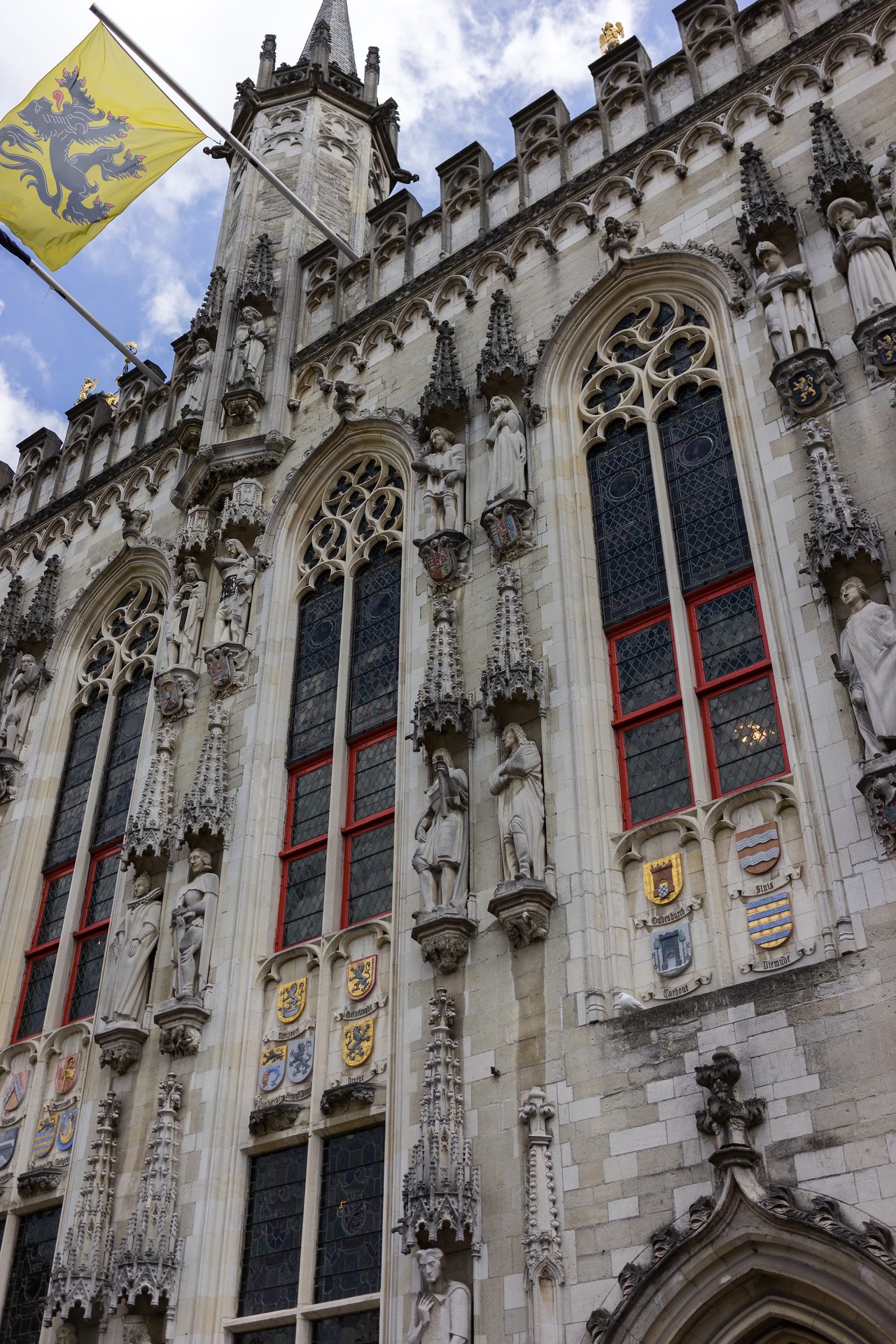 The facade of Bruges Town Hall with tall windows, detailed stone carvings, statues, and a flag waving above.