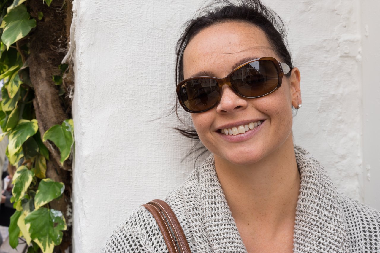 A woman wearing sunglasses and a knitted sweater smiles while leaning against a white wall.