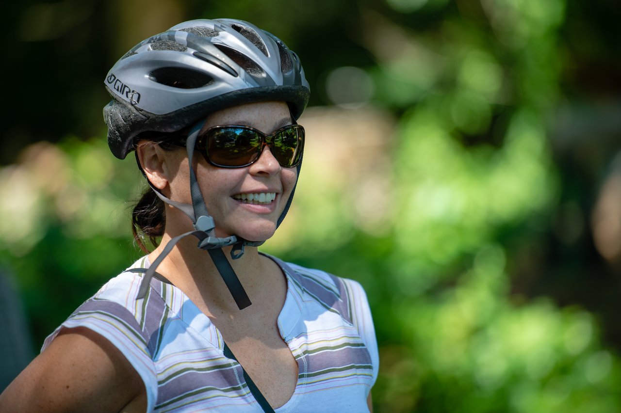 A woman wearing a bike helmet and sunglasses smiles while enjoying a bike ride outdoors.