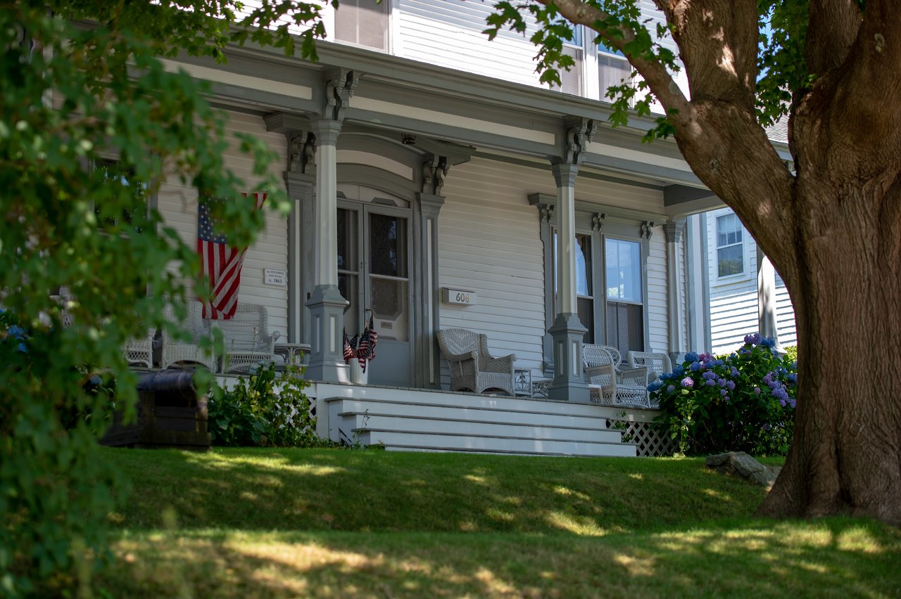 A white house with a porch, wicker chairs, and American flags, partially framed by trees and greenery.