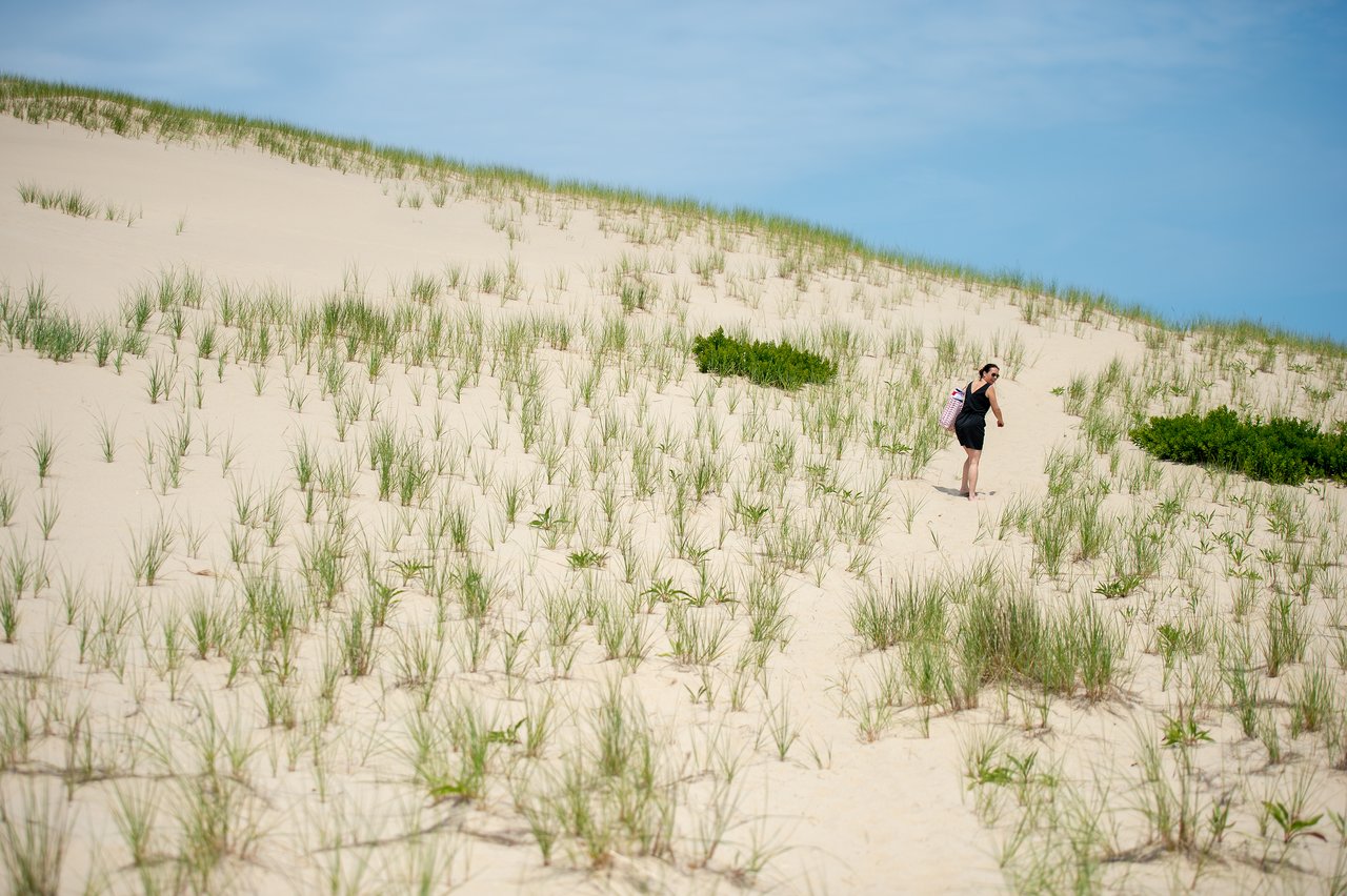 A person with a backpack walks up a sandy trail surrounded by grass and dunes under a clear sky.