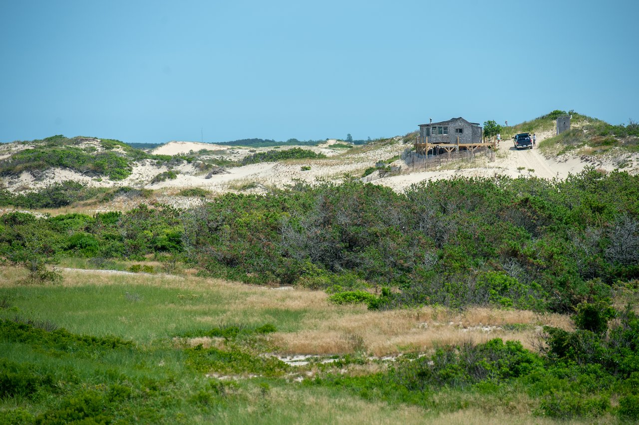 A sandy trail leads to a wooden shack on a dune, with a vehicle parked nearby.