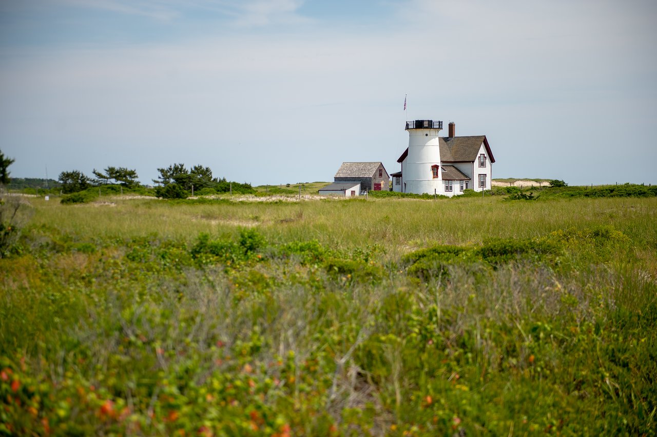 A white lighthouse with an attached house stands in a grassy field under a clear sky.