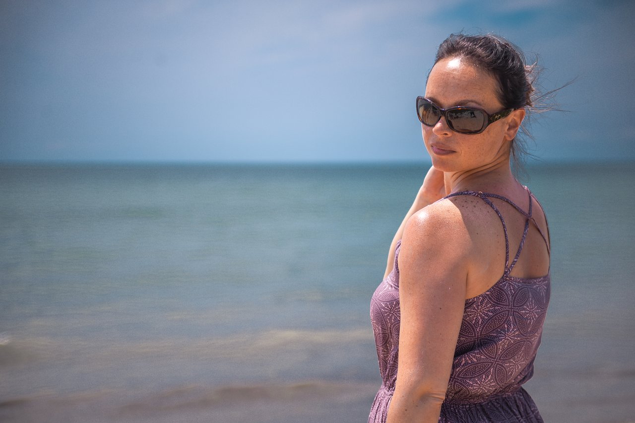 A woman wearing sunglasses and a patterned dress stands on the beach, looking back toward the camera.
