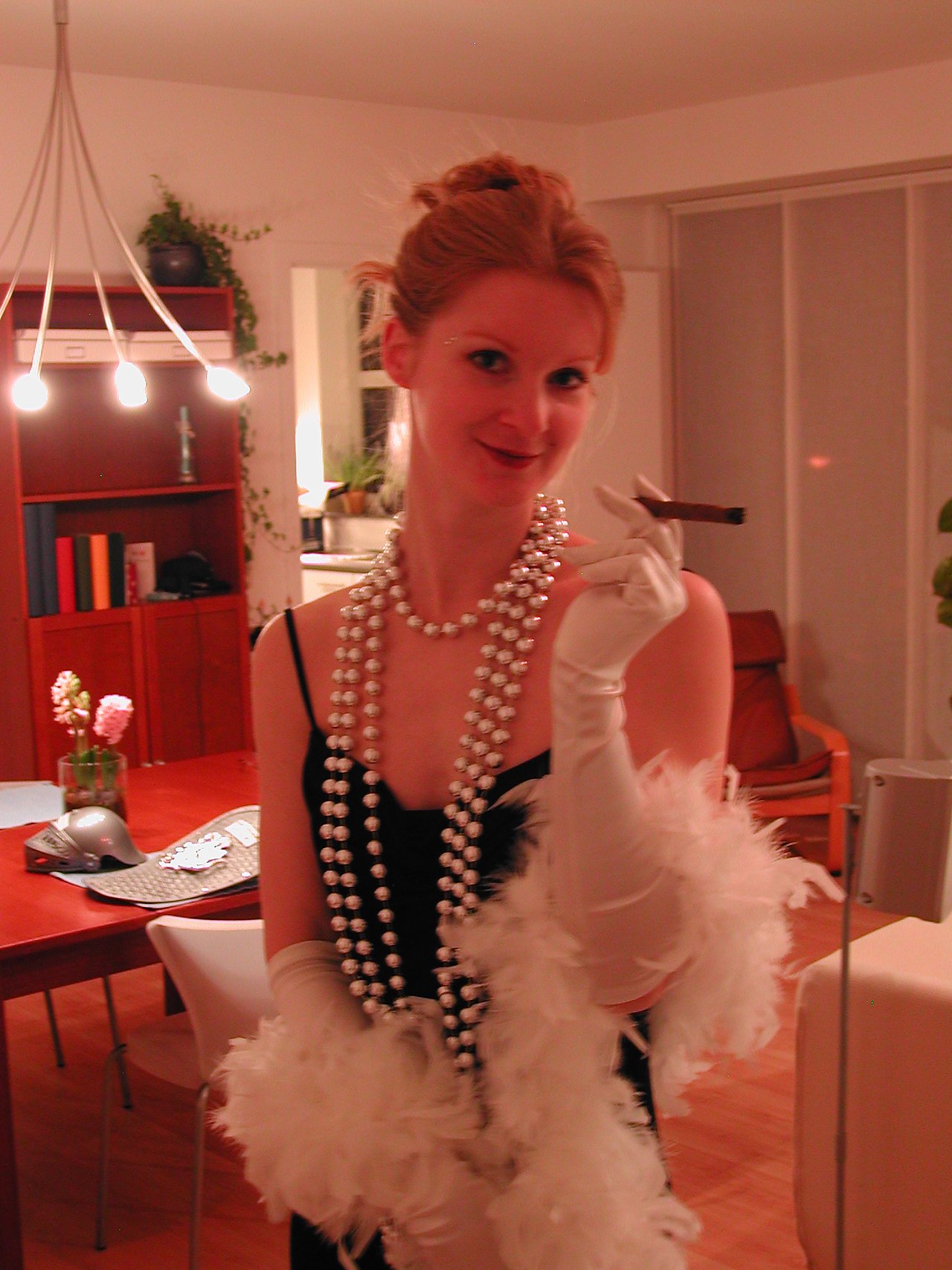 A woman in a glamorous costume with pearls, gloves, and a feather boa holds a cigar and smiles.