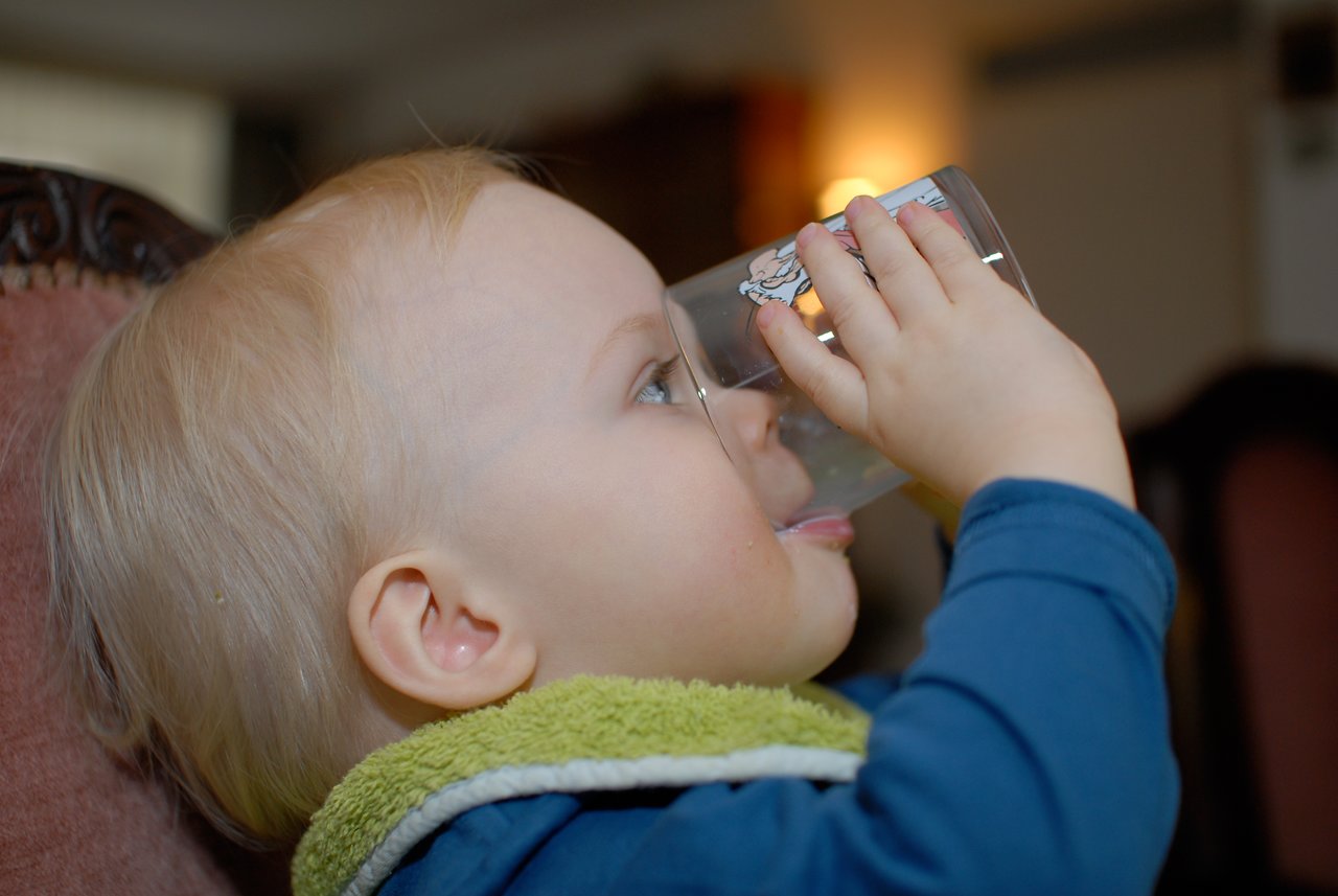 A young child in a blue outfit drinks from a glass, tilting it while sticking out their tongue.