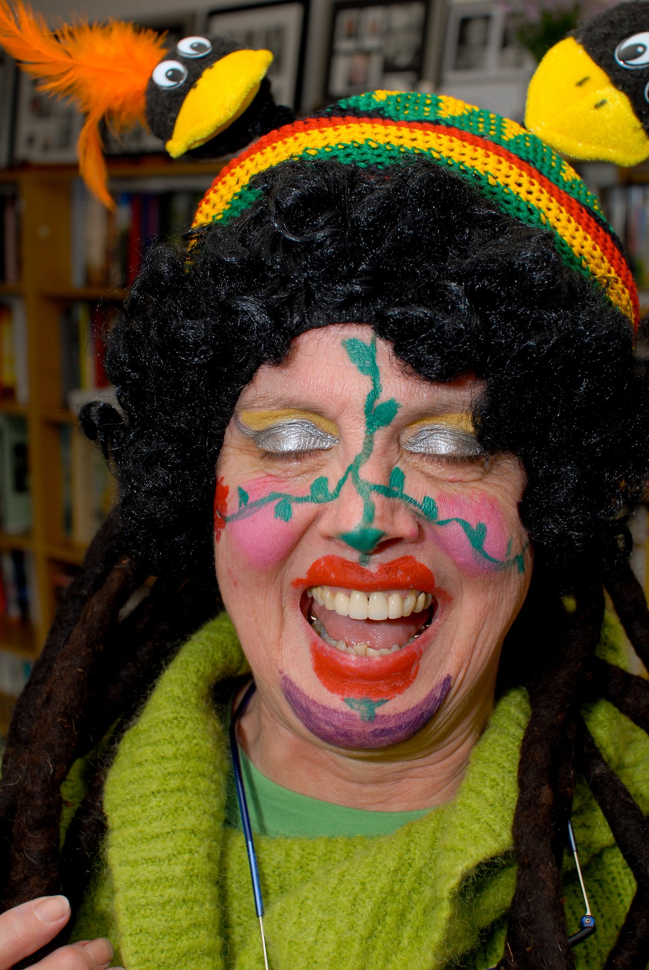 Person in a colorful costume with face paint, a curly wig, and a festive hat, smiling widely at the camera.