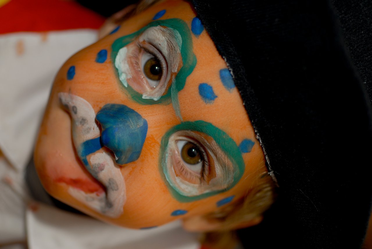Child with colorful face paint and a black hat looks directly at the camera during a carnival celebration.