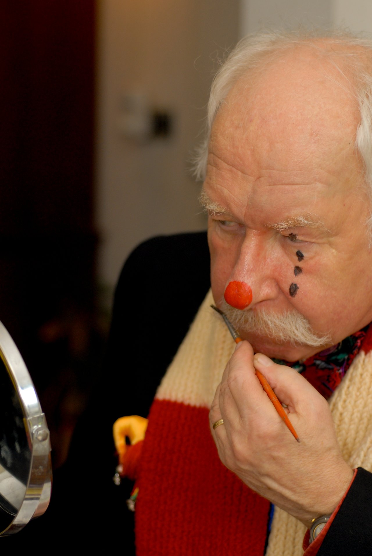 An older man paints his face like a clown, adding details near his eye while looking into a mirror.
