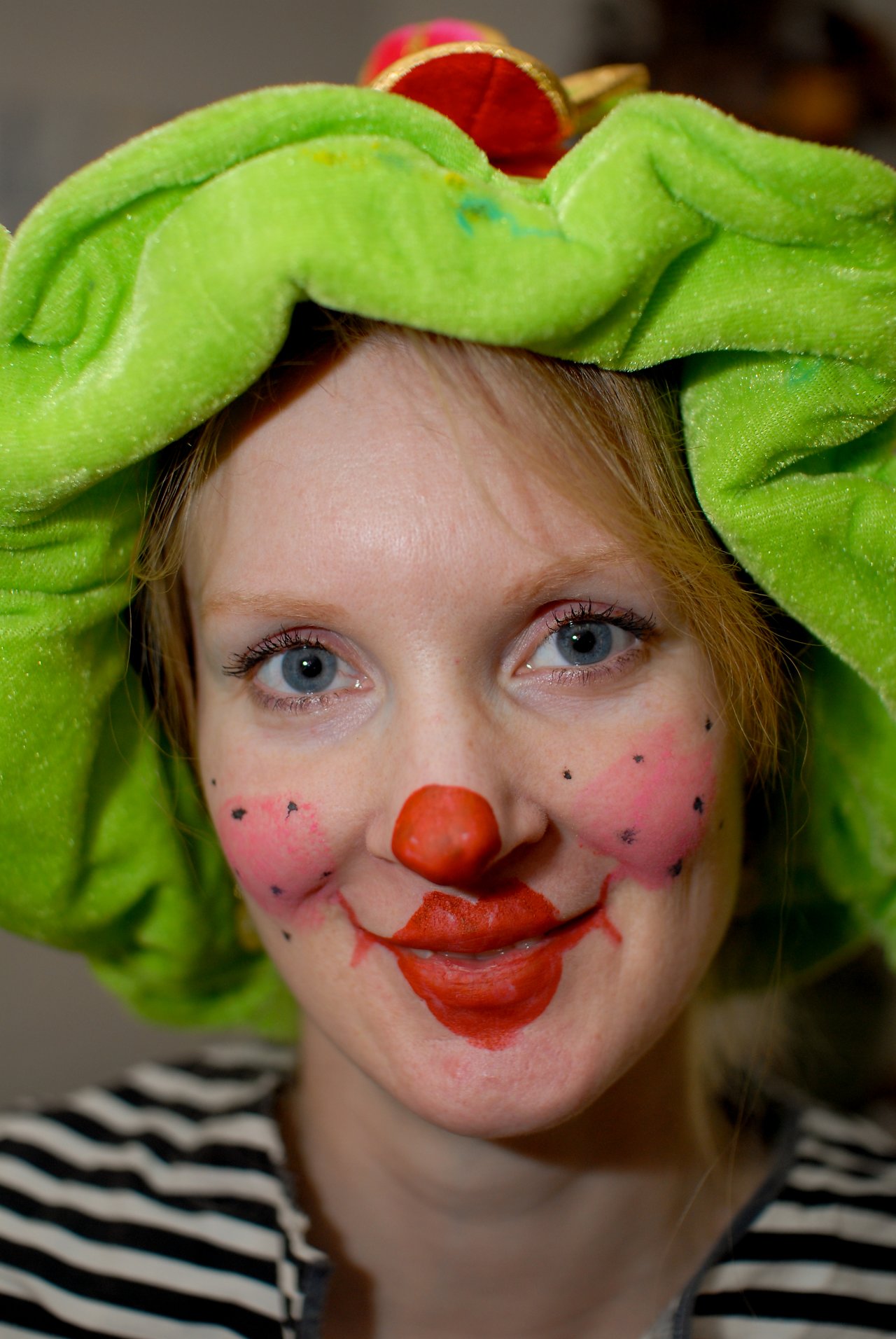Person dressed as a clown with face paint, a red nose, and a green plush hat, smiling at the camera.